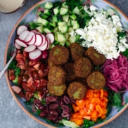 A large bowl of grand falafel salad plant-based entree for lunch or dinner with radishes, olives, peppers, cucumbers, cherry tomatoes, pickled pink onions, and vegan or dairy crumbled feta on a bed of massaged kale and iceburg lettuce