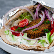 A large pita bread filled with plant-based falafel and tahini dressing or dairy-free tzatziki sauce and topped with onion, lettuce, tomato, and fresh dill