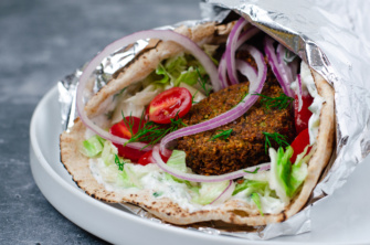 A large pita bread filled with plant-based falafel and tahini dressing or dairy-free tzatziki sauce and topped with onion, lettuce, tomato, and fresh dill