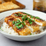 A bowl of rice topped with plant-based panko-crusted planetarian "chicken" cutlets made from tofu and topped with katsu-style sauce and herbs