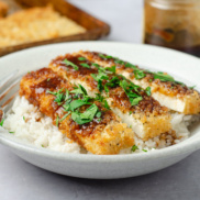 A bowl of rice topped with plant-based panko-crusted planetarian "chicken" cutlets made from tofu and topped with katsu-style sauce and herbs