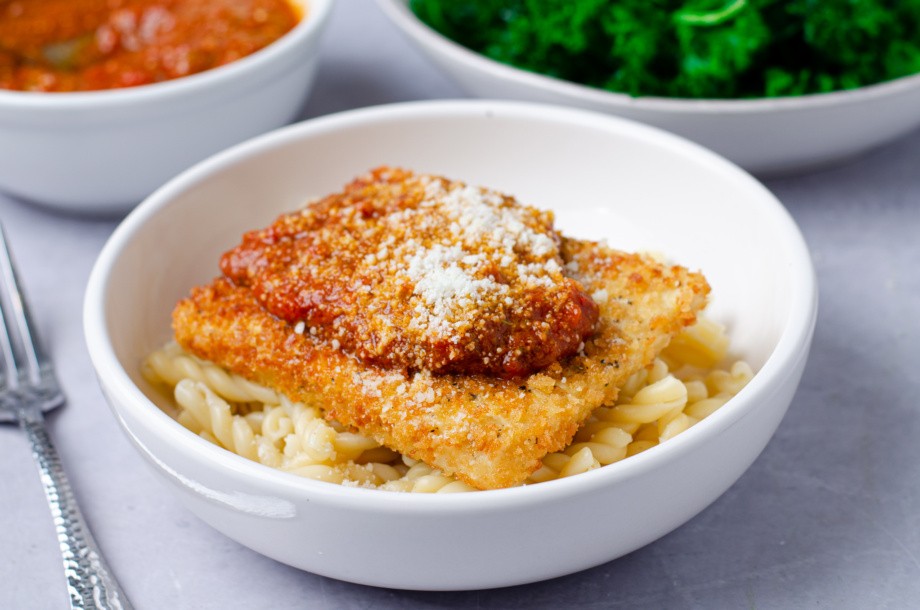 A bowl of pasta topped with plant-based panko-crusted planetarian "chicken" cutlets made from tofu and topped with pesto flavored tomato sauce and parmesan cheese. You won't know this chicken parm is vegan