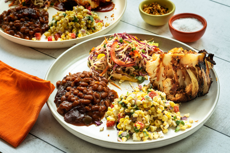 A plate full of classic summer staples such as corn salad, baked beans, coleslaw, and barbecue made plant-based