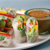 A platter of freshly made plant-based, gluten free, spring rolls with peppers, cucumbers, radish, vermicelli rice noodles, and avocado wrapped in rice paper and served with a side of peanut sauce
