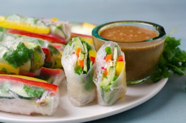 A platter of freshly made plant-based, gluten free, spring rolls with peppers, cucumbers, radish, vermicelli rice noodles, and avocado wrapped in rice paper and served with a side of peanut sauce