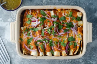 A baking dish of plant-based enchiladas with beans, dairy-free cheese, frozen spinach, salsa, and topped with red onion and cilantro